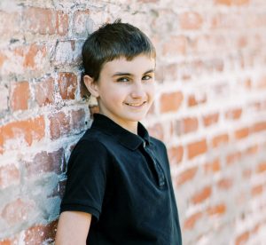 National Foster Care Month | little boy who is waiting to be adopted smiling against brick wall 