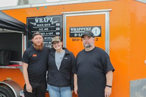 Wrapperz D-Lite | Richmond VA Food Truck | Owners standing in front of their bright orange food truck smiling