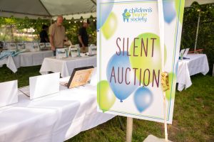 A blue and lime green sign with balloons on it that reads "Silent Auction" in front of a long table of auction items.