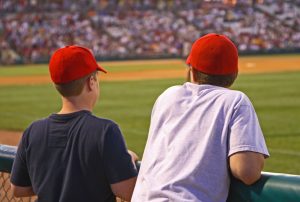 two younger boys watching a baseball game from the stands. They are wearing red baseball caps.