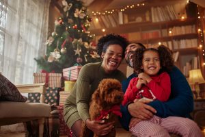 A happy diverse family all laughing and smiling sitting on their floor with their dog around their Christmas tree. There are wrapped presents around the tree.