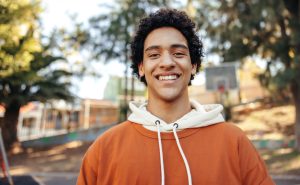 A teenage boy smiling wearing a orange and white hoodie. He is standing in front of his school. He has black curly hair.