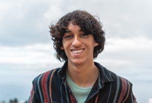 Portrait of happy young teenager smiling in front of the camera. He has long, curly dark brown hair and is wearing a striped collared shirt.