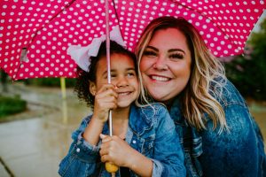 A young mom smiling under a pink polka dot umbrella with her adopted daughter.