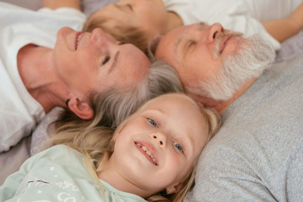 two little children with their grandparents. They are all lying on the floor and their heads are touching. They are all smiling.