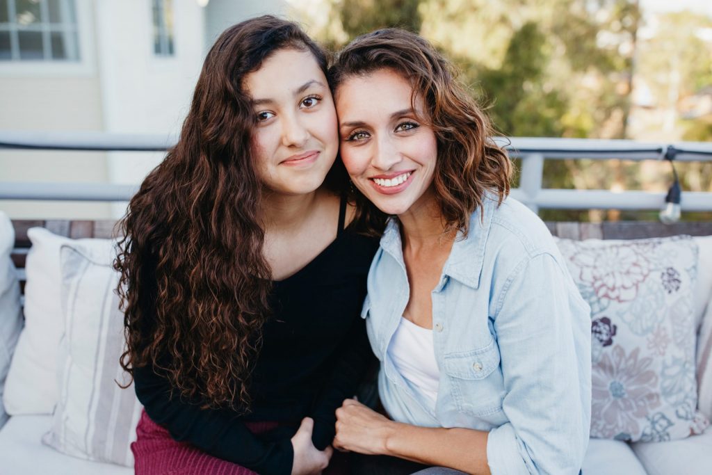 An adoptive mother with her adopted daughter. They are on an outdoor couch smiling and holding hands.