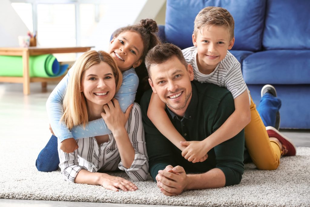 Happy couple, man and woman, smiling with their adopted children, one boy and one girl, who are both leaning on their parents' backs.