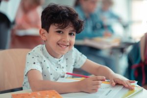 A young boy in elementary school sitting at his desk, smiling. He is working on his homework.