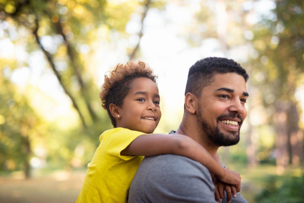 An African American son and dad. The dad is giving his son a piggy-back ride. They are both smiling. There are green trees behind them. This image represents National Foster Care Month. Would you consider adopting from foster care?