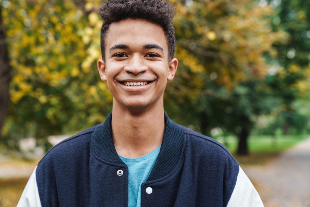 A teenage boy wearing a letterman's jacket smiling at the camera in front of changing trees in the fall.